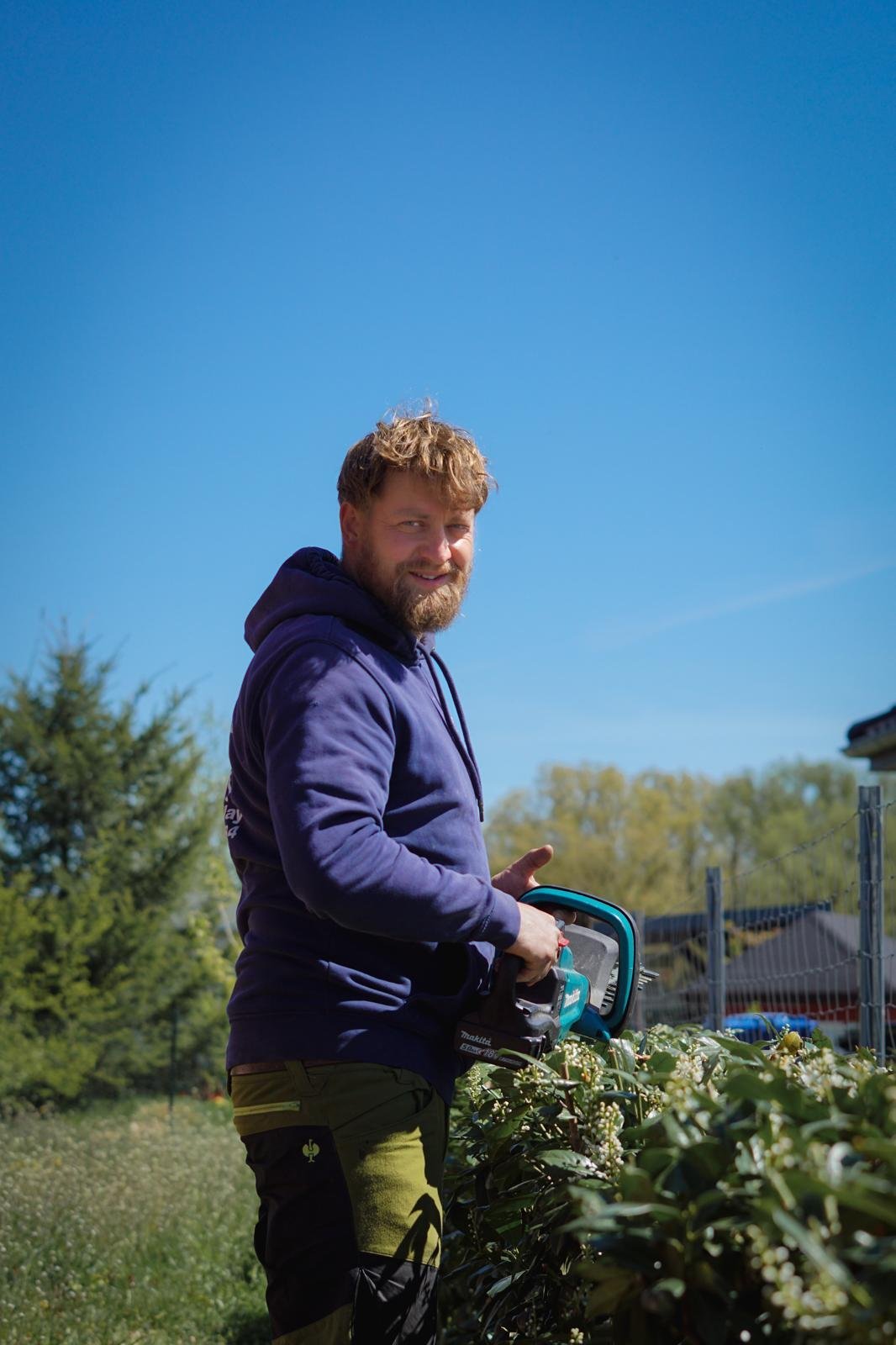 Man with reddish-blond beard wearing a navy hoodie holding garden shears outdoors with clear blue sky and green landscape behind him