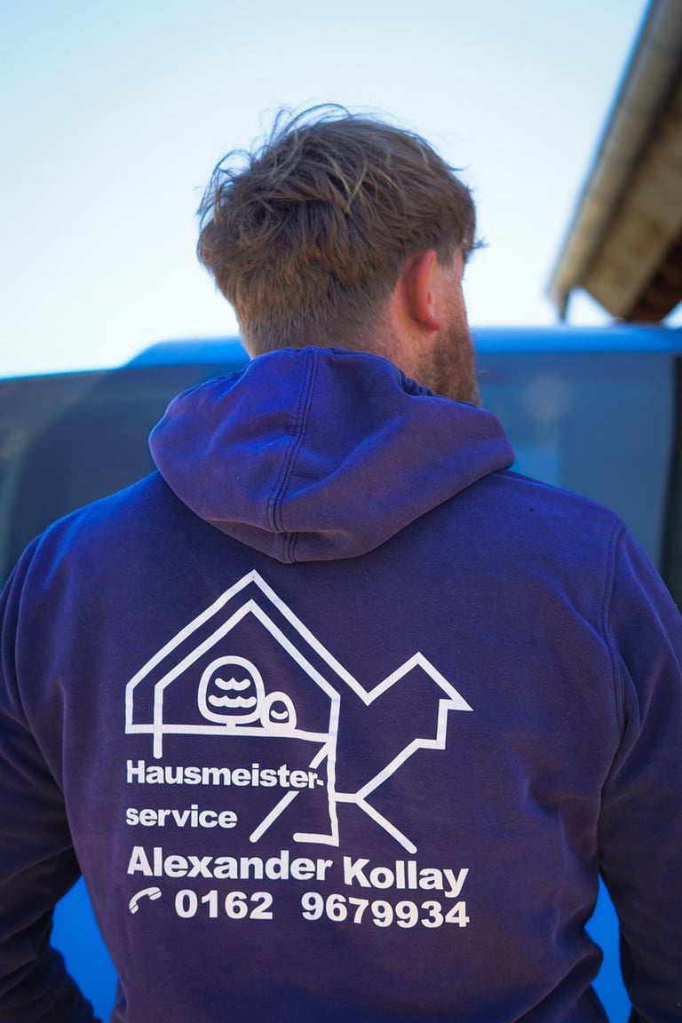 Man wearing a navy blue hoodie with Hausmeister service branding and contact details, viewed from behind against a light blue sky